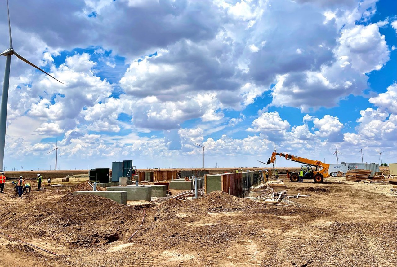 Construction site with wind turbines in the background, workers and machinery under a blue sky.