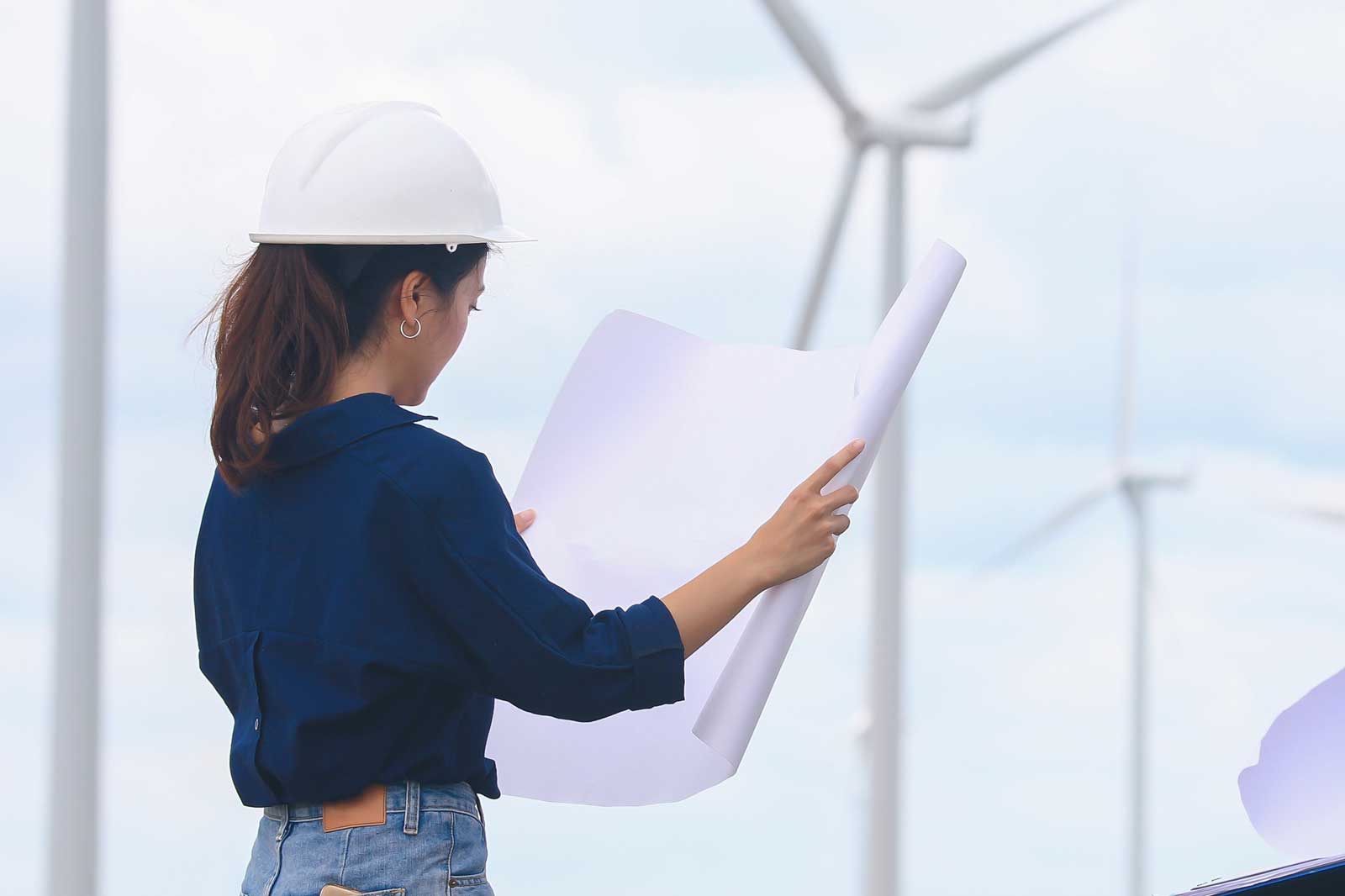 A person in a white hard hat holds blueprints and stands near wind turbines under a cloudy sky.