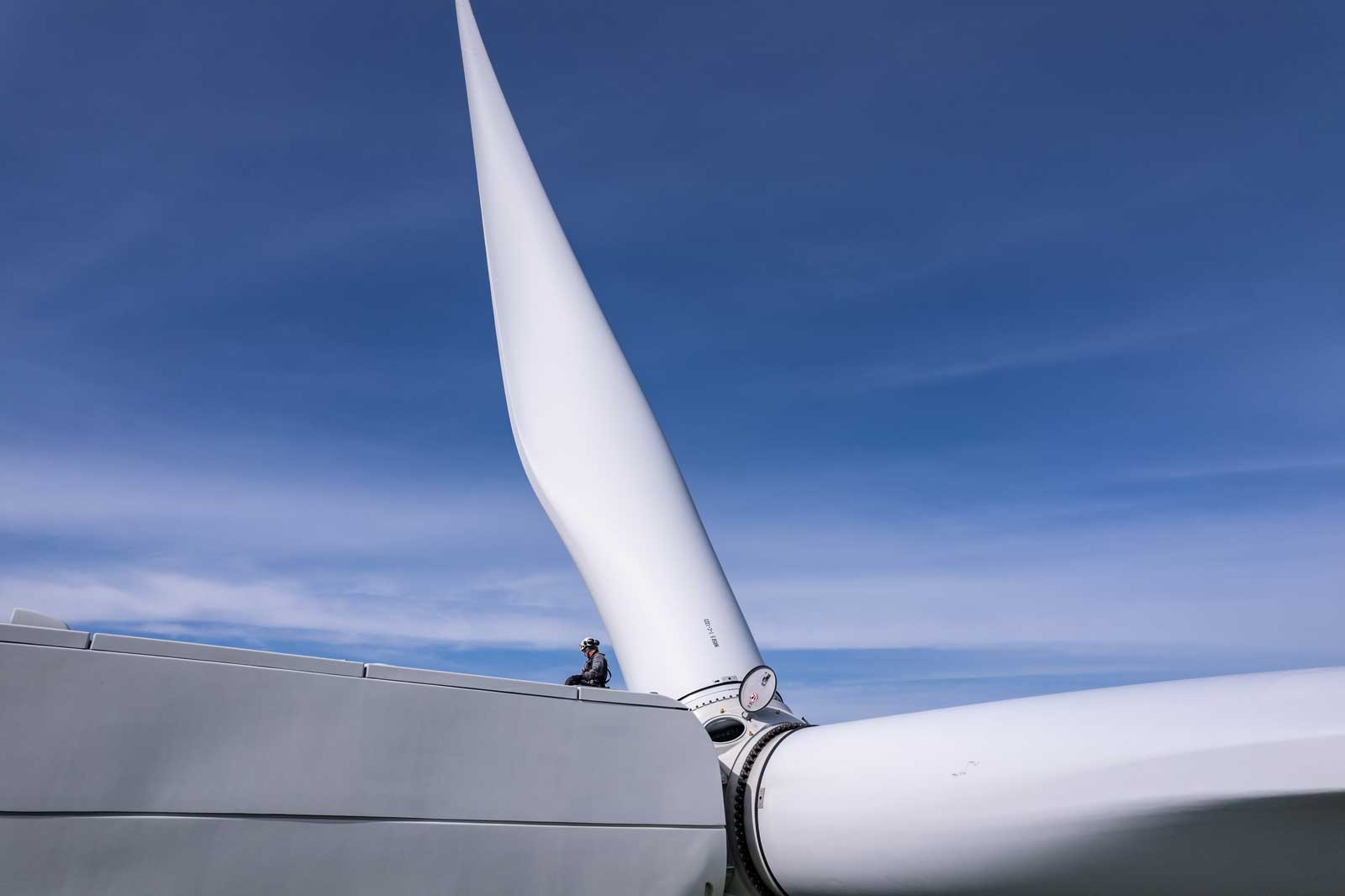 A person sits atop a wind turbine, beside a large white blade, with a clear blue sky in the background.