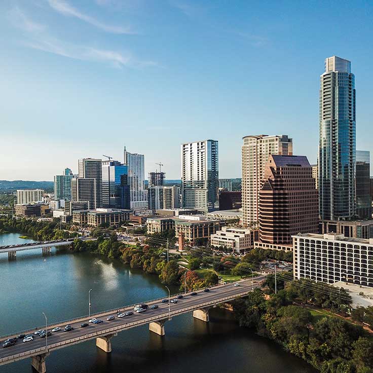 Aerial view of a city skyline beside a river, showcasing modern skyscrapers and a bridge with cars.