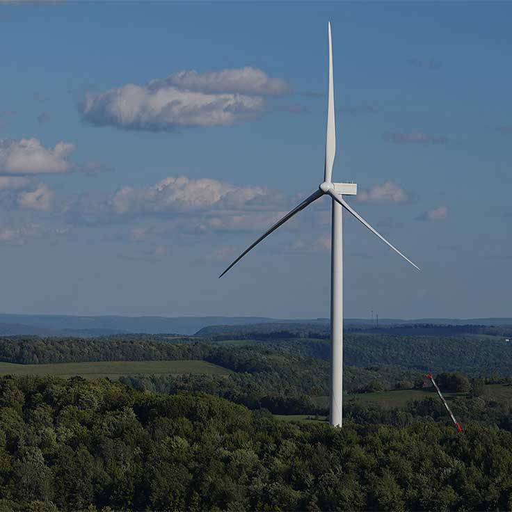 A tall wind turbine stands amidst lush green hills under a blue sky with scattered clouds.