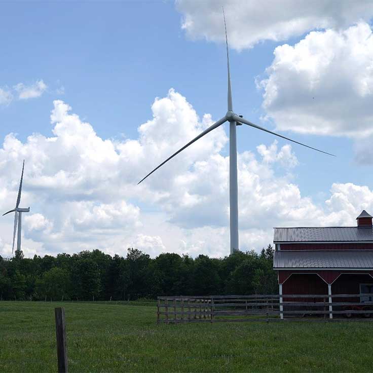Two large wind turbines stand in a field near a rustic barn under a bright blue sky with fluffy white clouds.