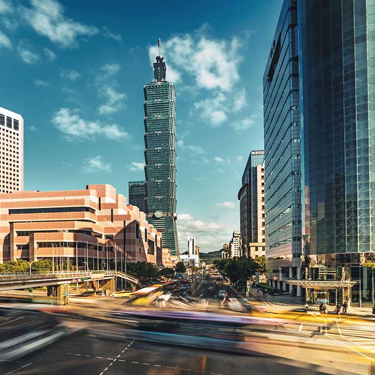 A bustling urban scene featuring Taipei 101 tower amidst modern skyscrapers under a clear blue sky with moving traffic below.