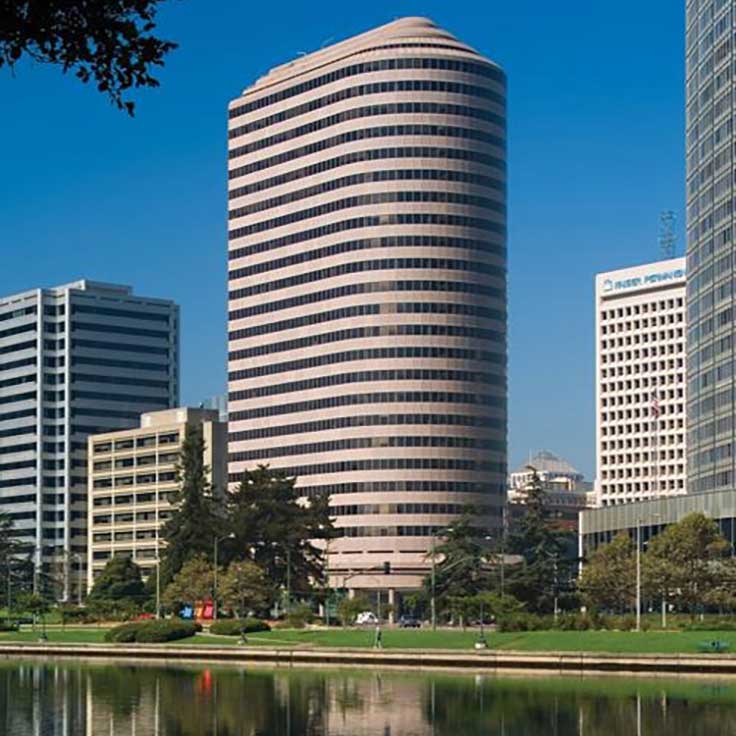 A modern building stands next to a park with trees and a pond, under a clear blue sky.