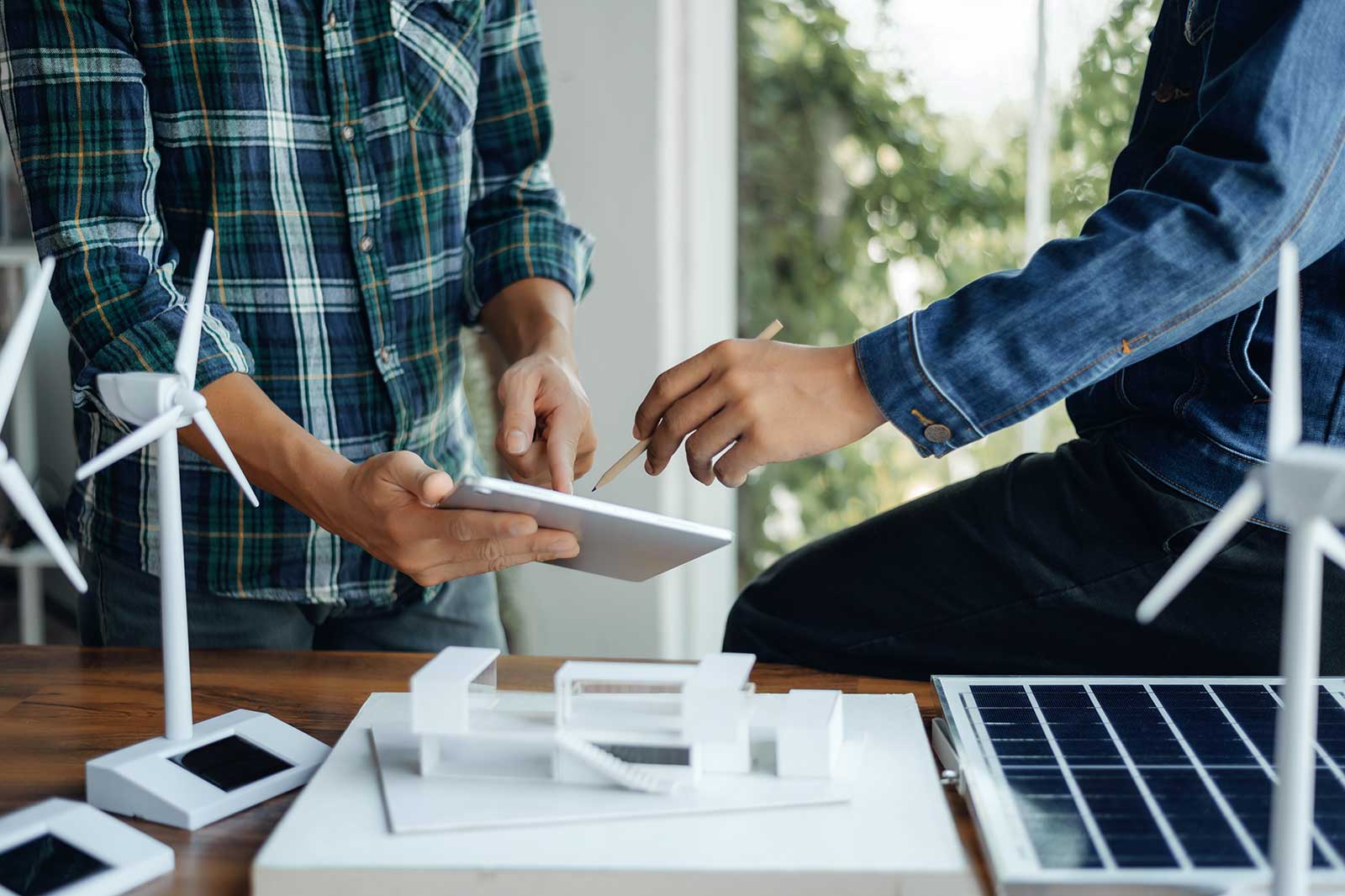 Two individuals collaborate on a tablet amidst models of wind turbines and solar panels.