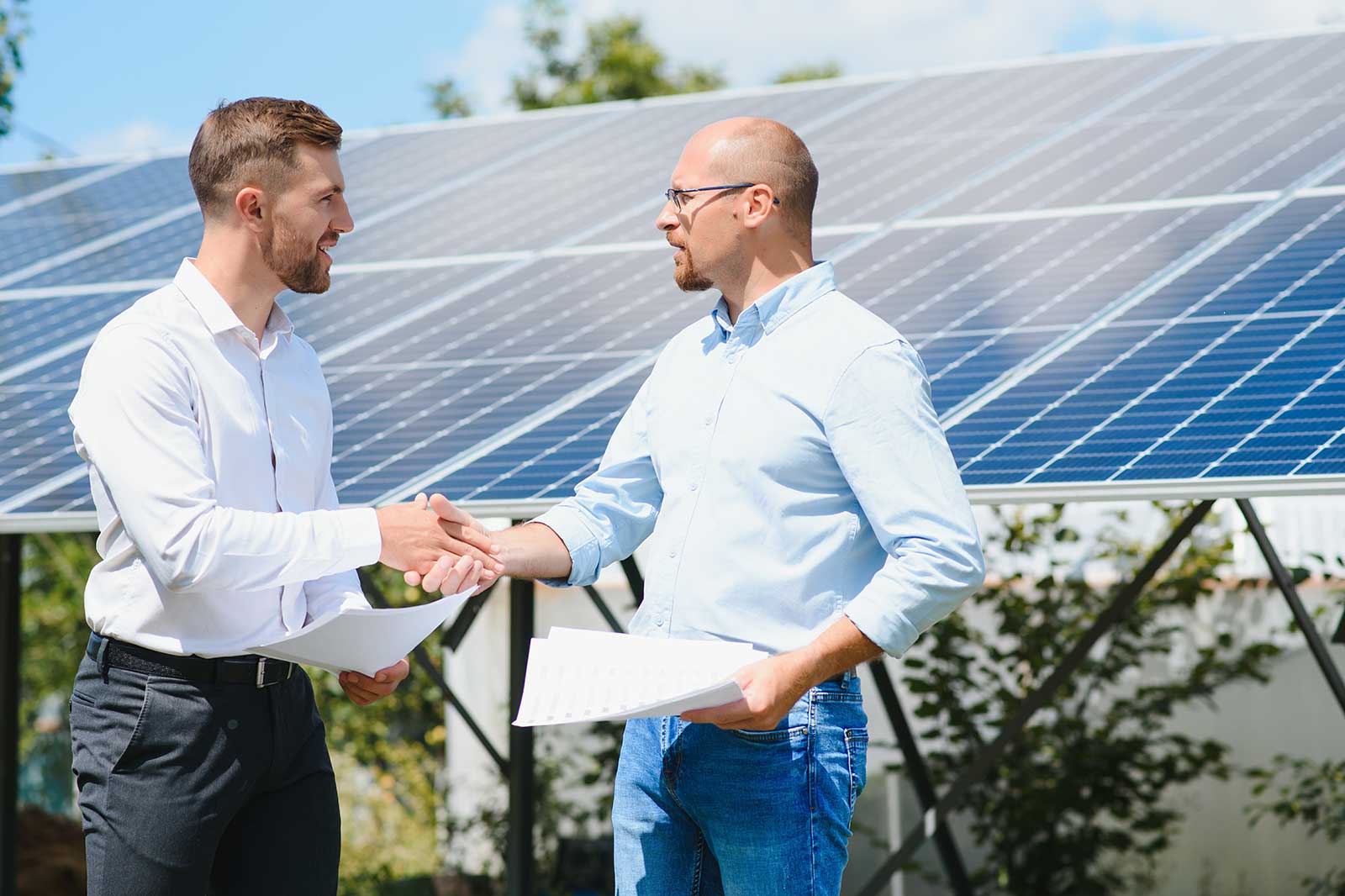 Two individuals shake hands in front of solar panels, symbolising a business agreement in renewable energy.