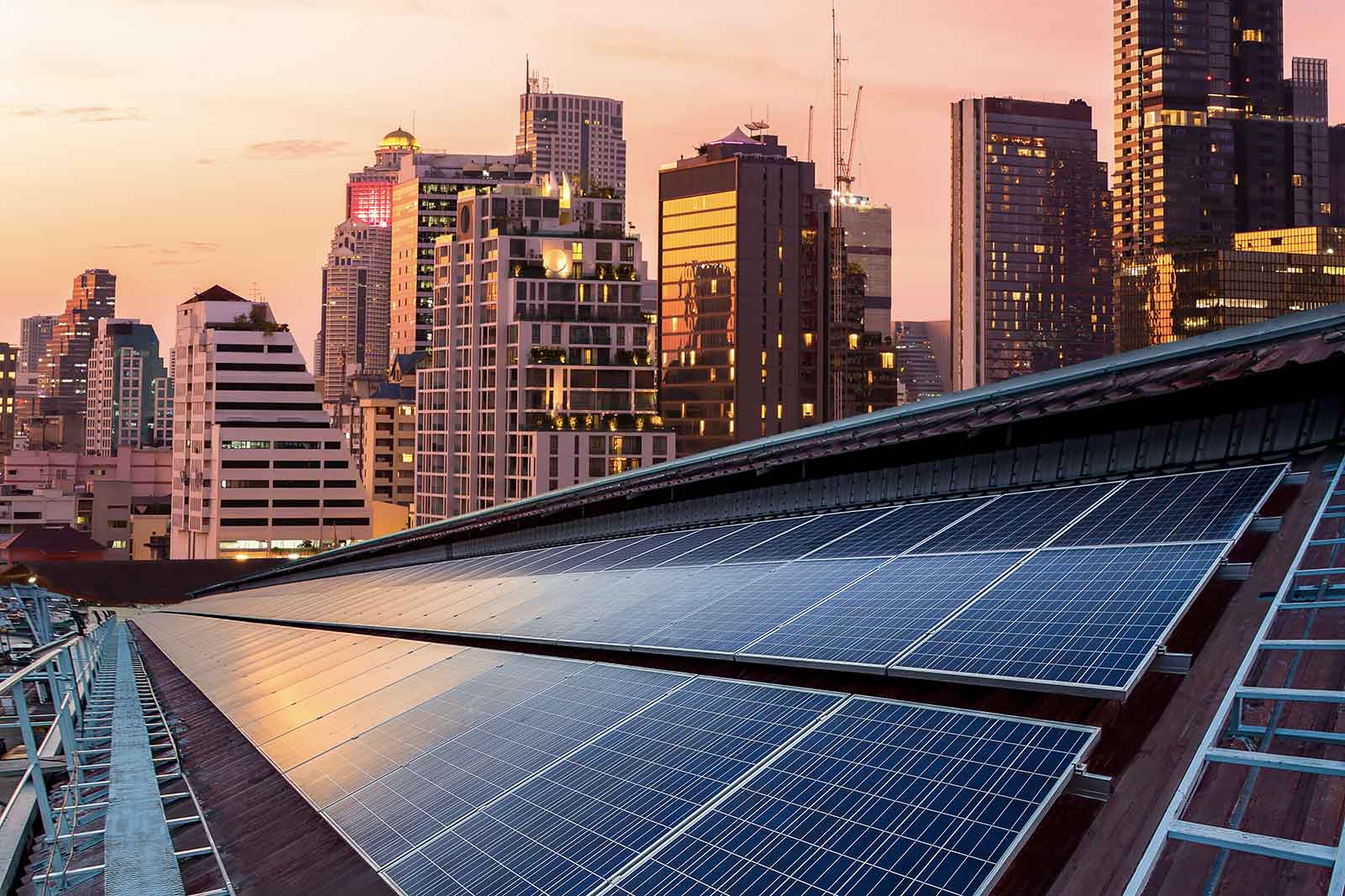 Solar panels on a rooftop with a city skyline at sunset, featuring modern skyscrapers and warm hues in the sky.
