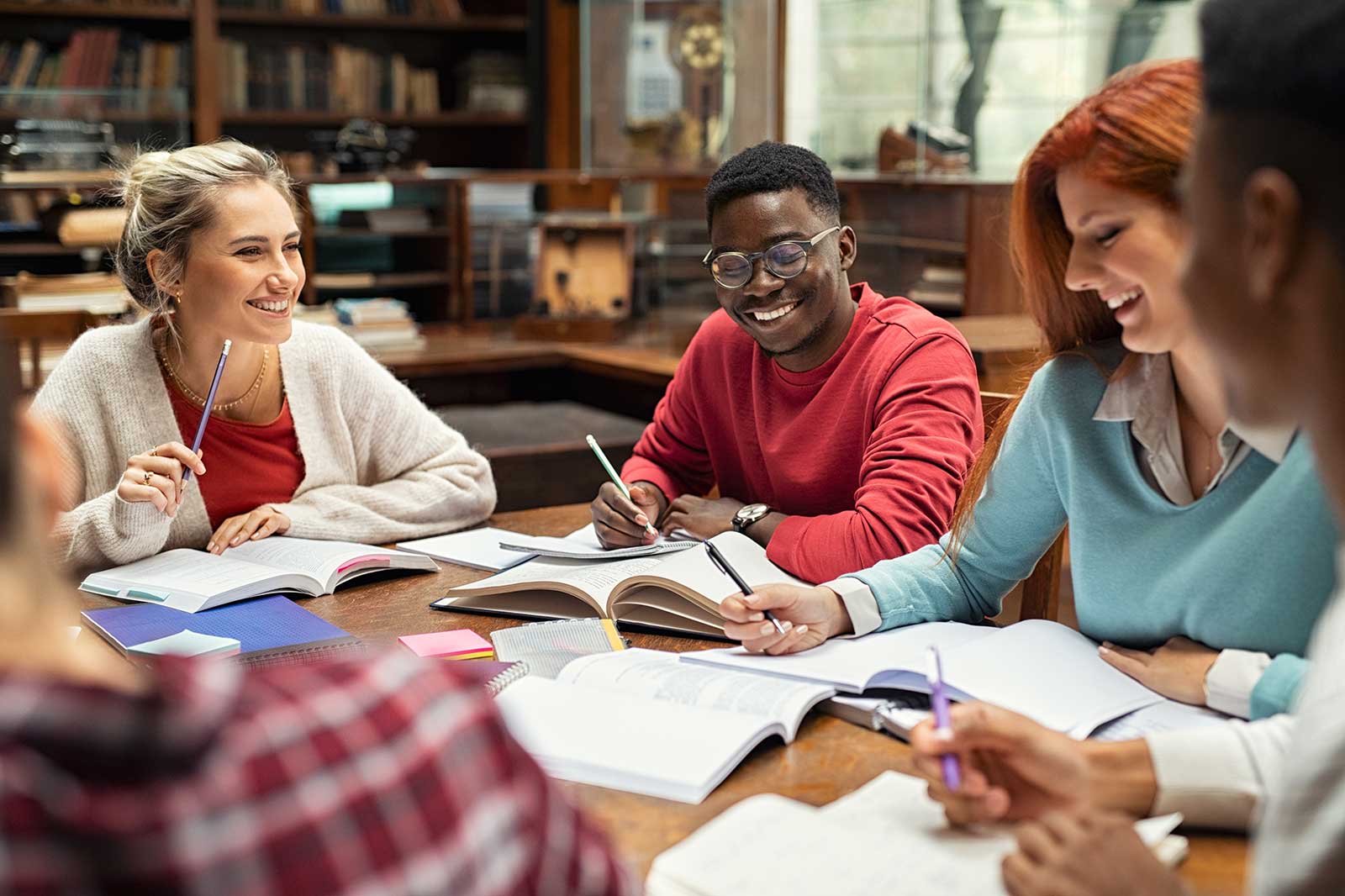 A group study session in a library with four individuals engaged in discussion and writing, surrounded by books and stationery.