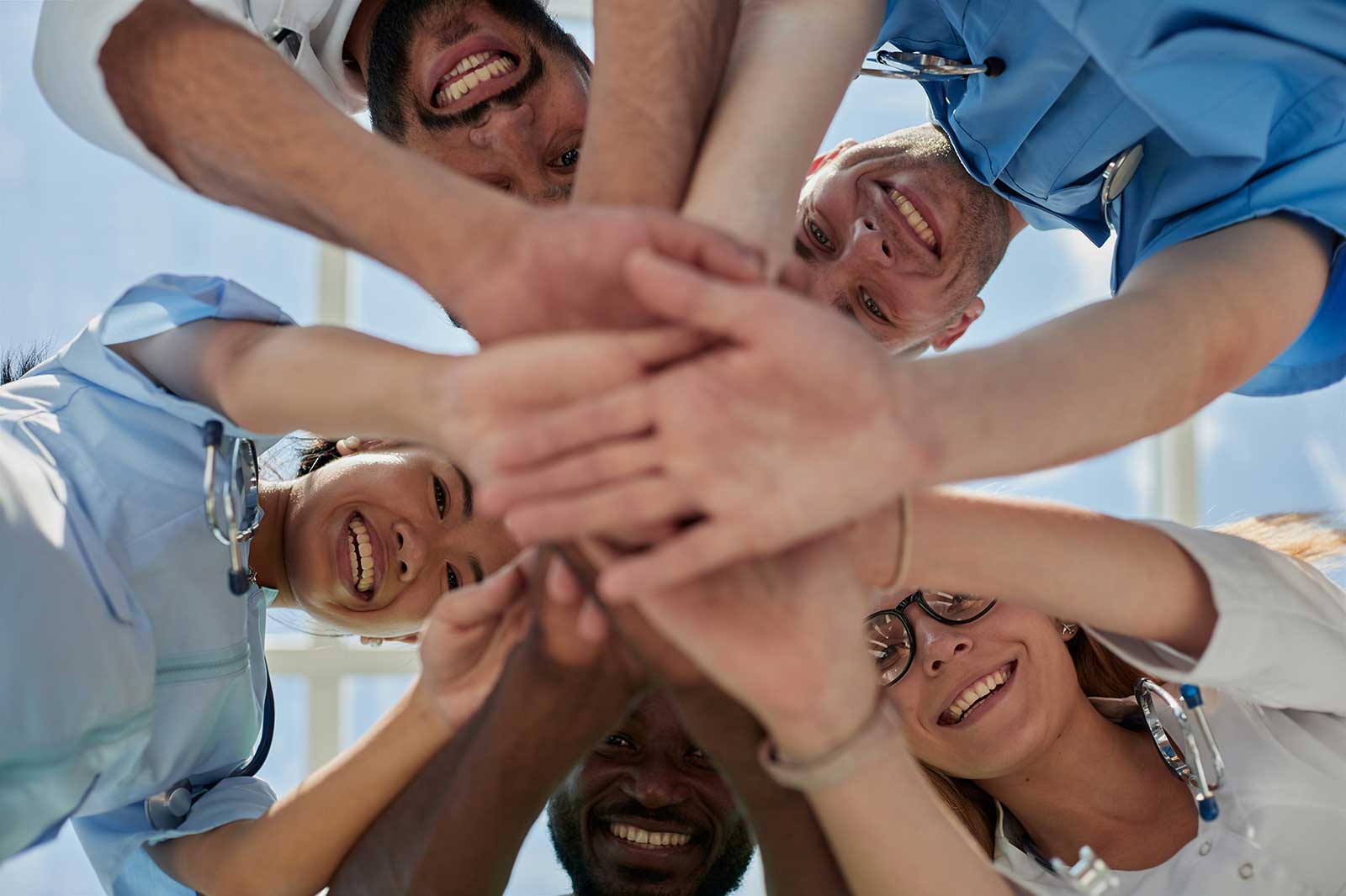 A group of healthcare professionals smiling and joining hands in a team spirit gesture in a bright environment.