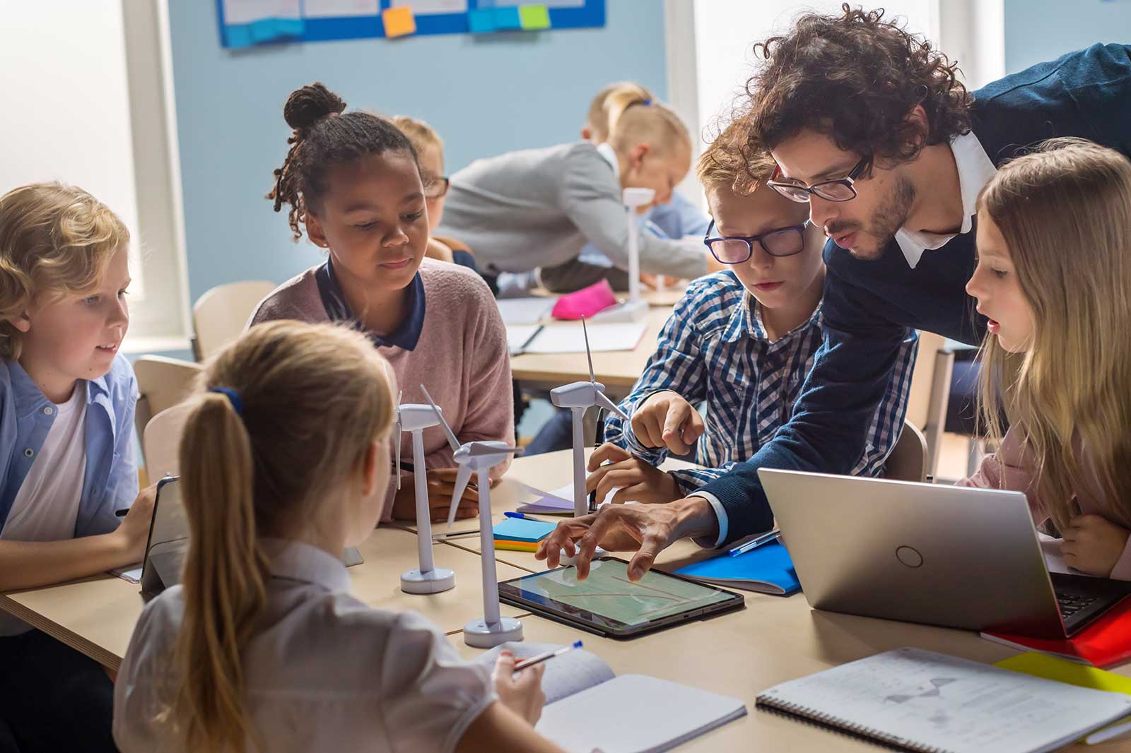 A teacher collaborates with students at a classroom table with laptops and wind turbine models, focusing on a tablet.