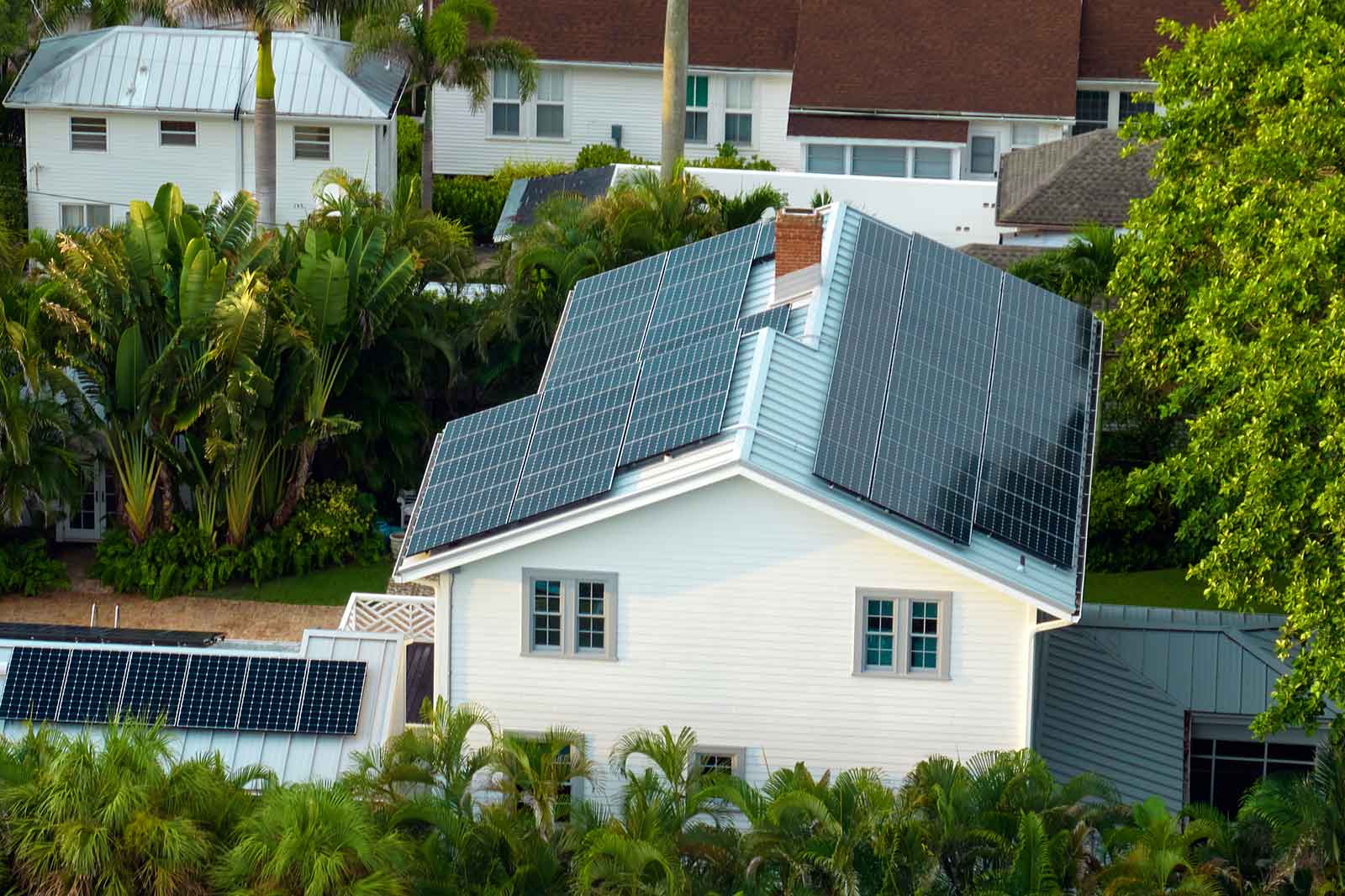 A white house with solar panels on the roof, surrounded by lush greenery and other homes in the background.