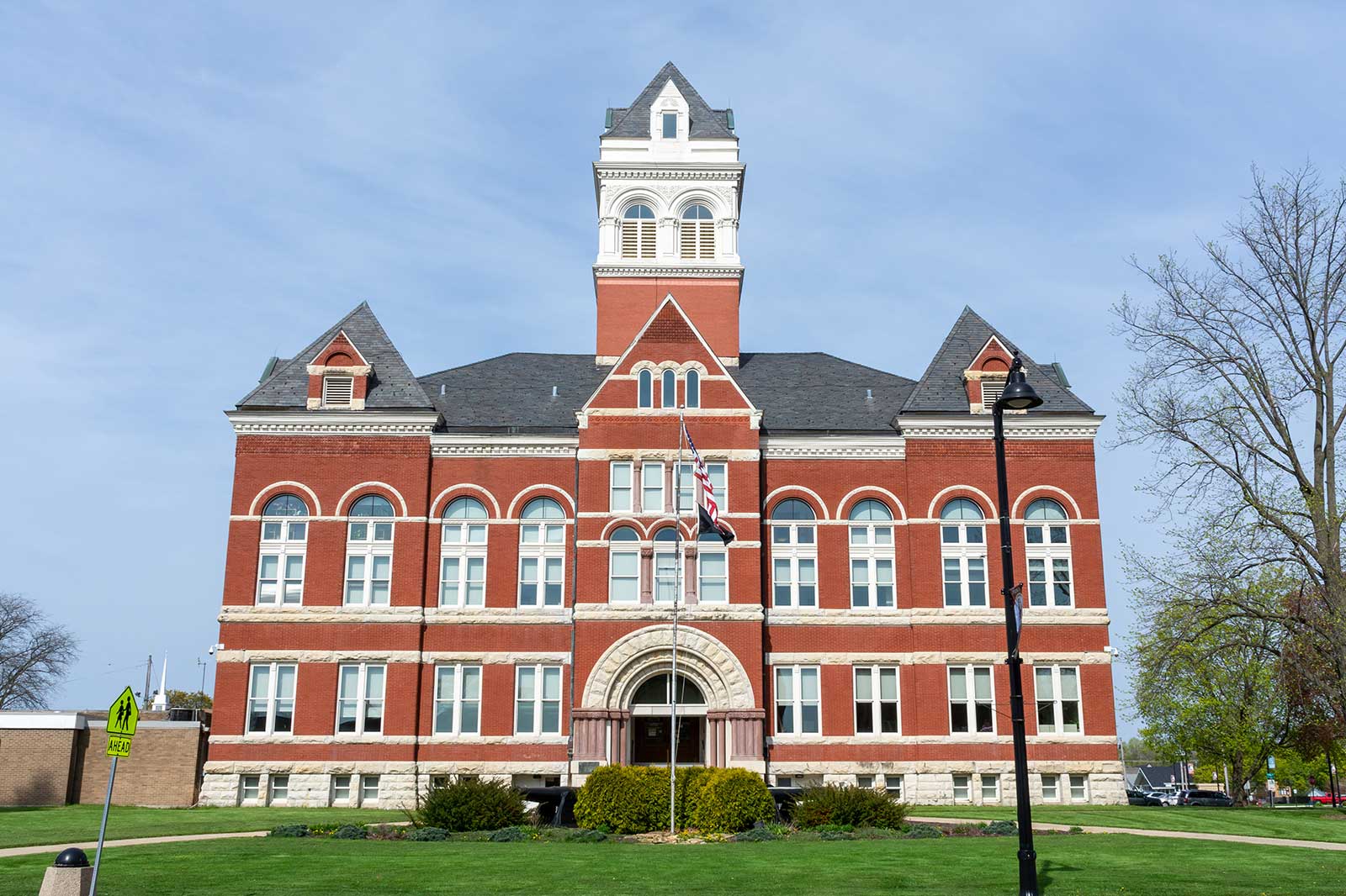 A historic red brick building with a tower and arched windows, set against a clear sky and greenery.