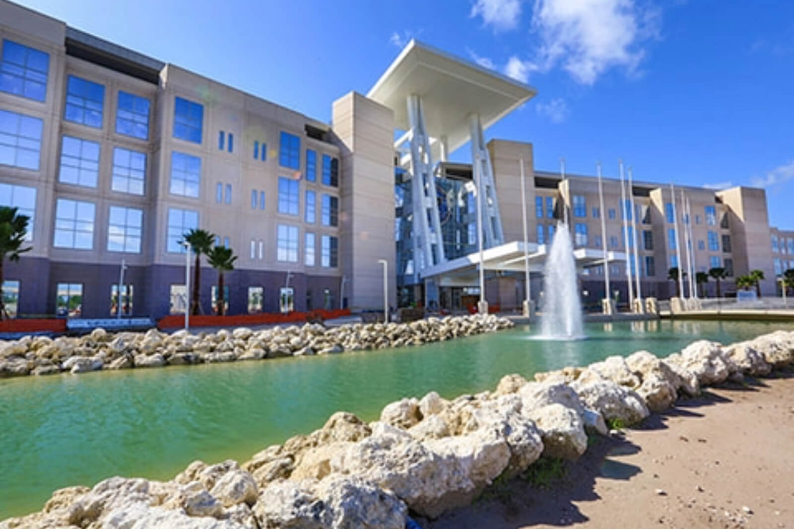 A modern building with large glass windows and a white roof structure, situated beside a green pond with a fountain.