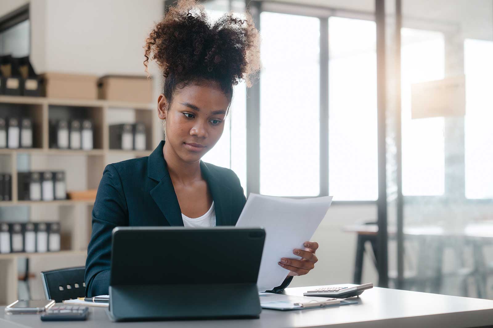 A person in a blazer holds documents while working at a desk with a tablet and calculator in a bright office space.