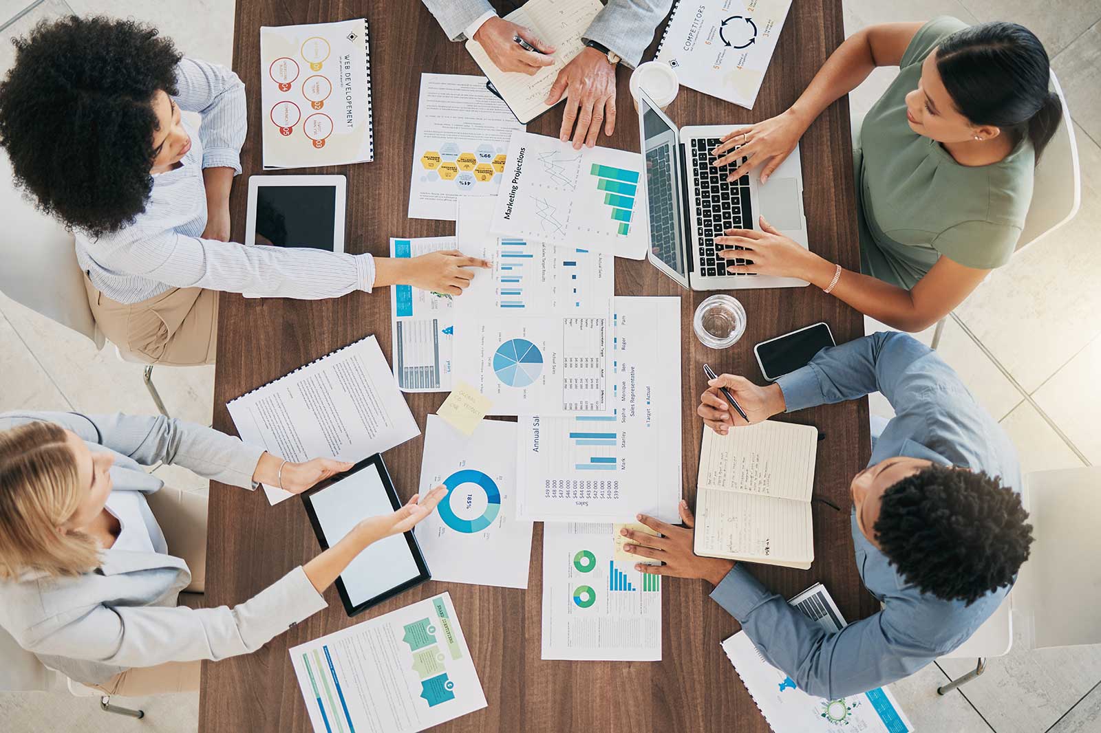 A diverse group collaborating on a business project, surrounded by charts, graphs, and electronic devices on a wooden table.
