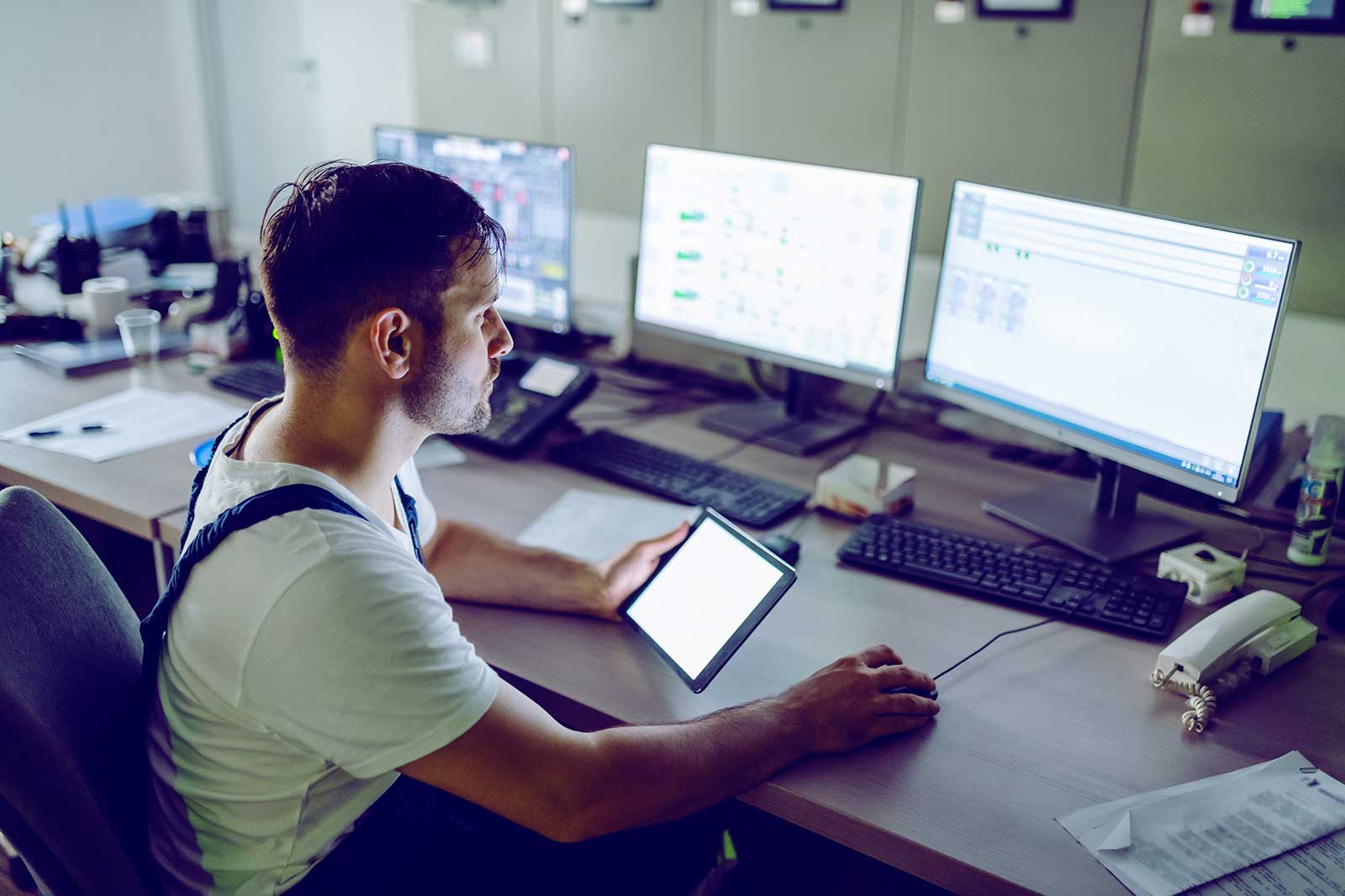 A user sits at a desk with multiple computer monitors, holding a tablet, surrounded by office equipment and documents.