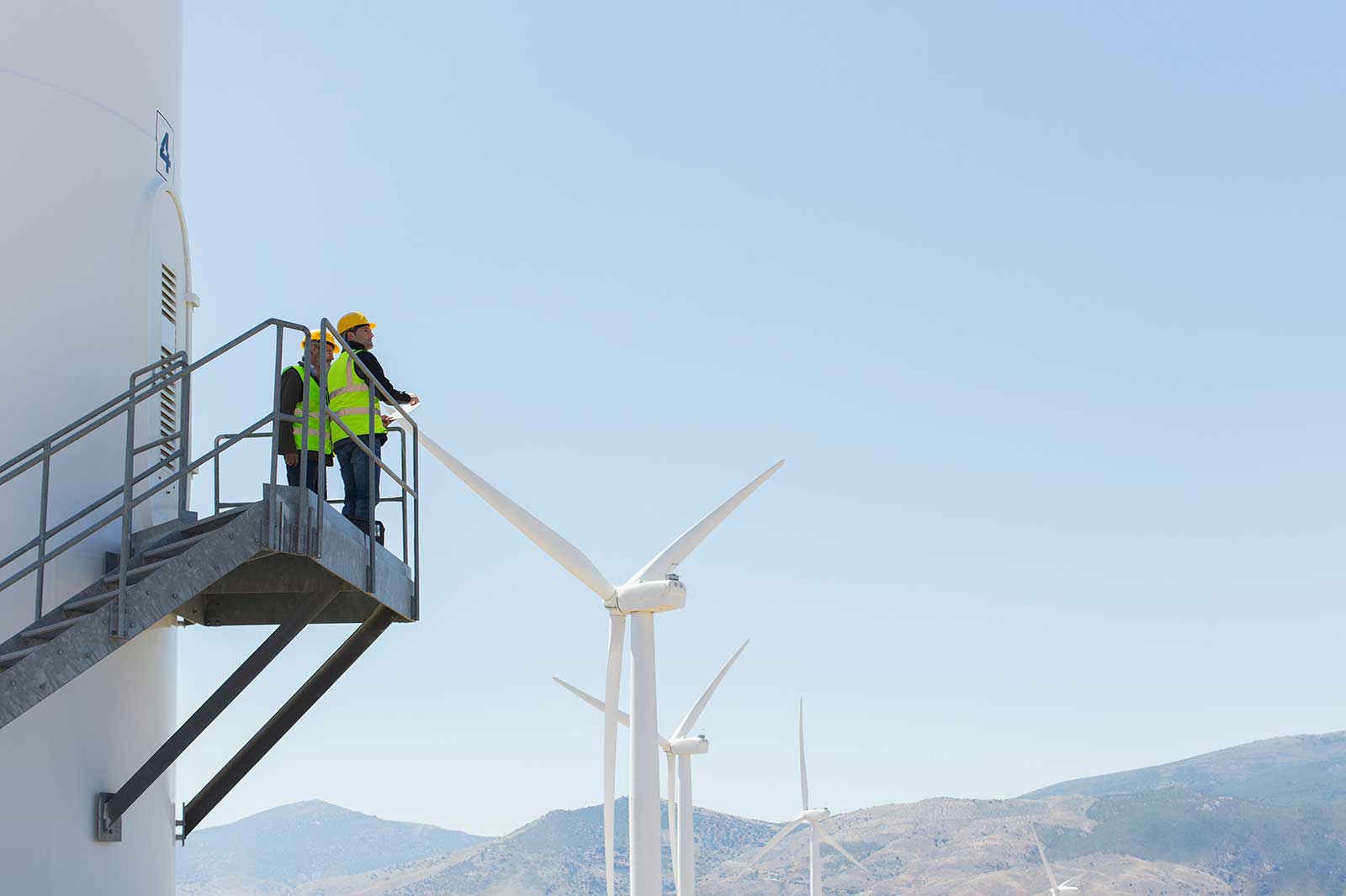 Two workers in safety gear assess a wind turbine from a platform, with multiple turbines visible under a clear blue sky.