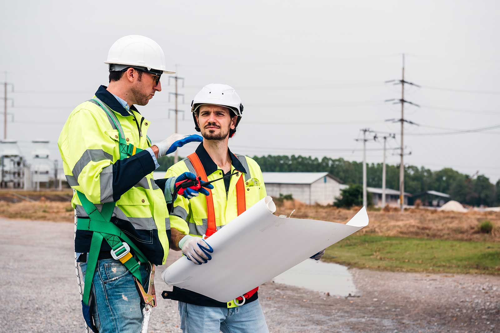 Two construction workers discuss plans on a site, one holding blueprints. They wear safety helmets and high-visibility jackets.