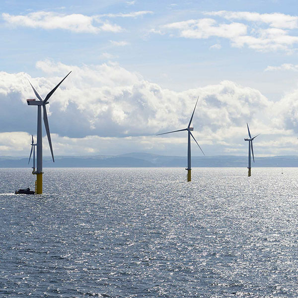 Three offshore wind turbines stand on yellow bases in a shimmering sea under a cloudy sky.