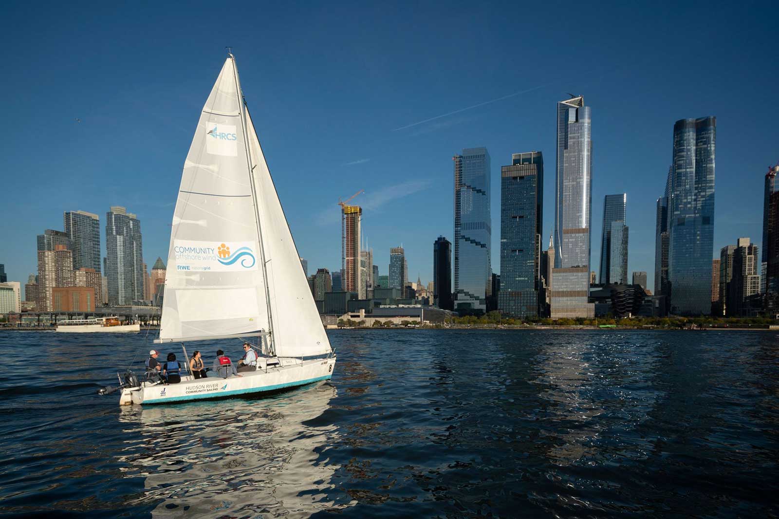 A sailing boat glides through calm waters with a backdrop of modern skyscrapers under a clear blue sky.