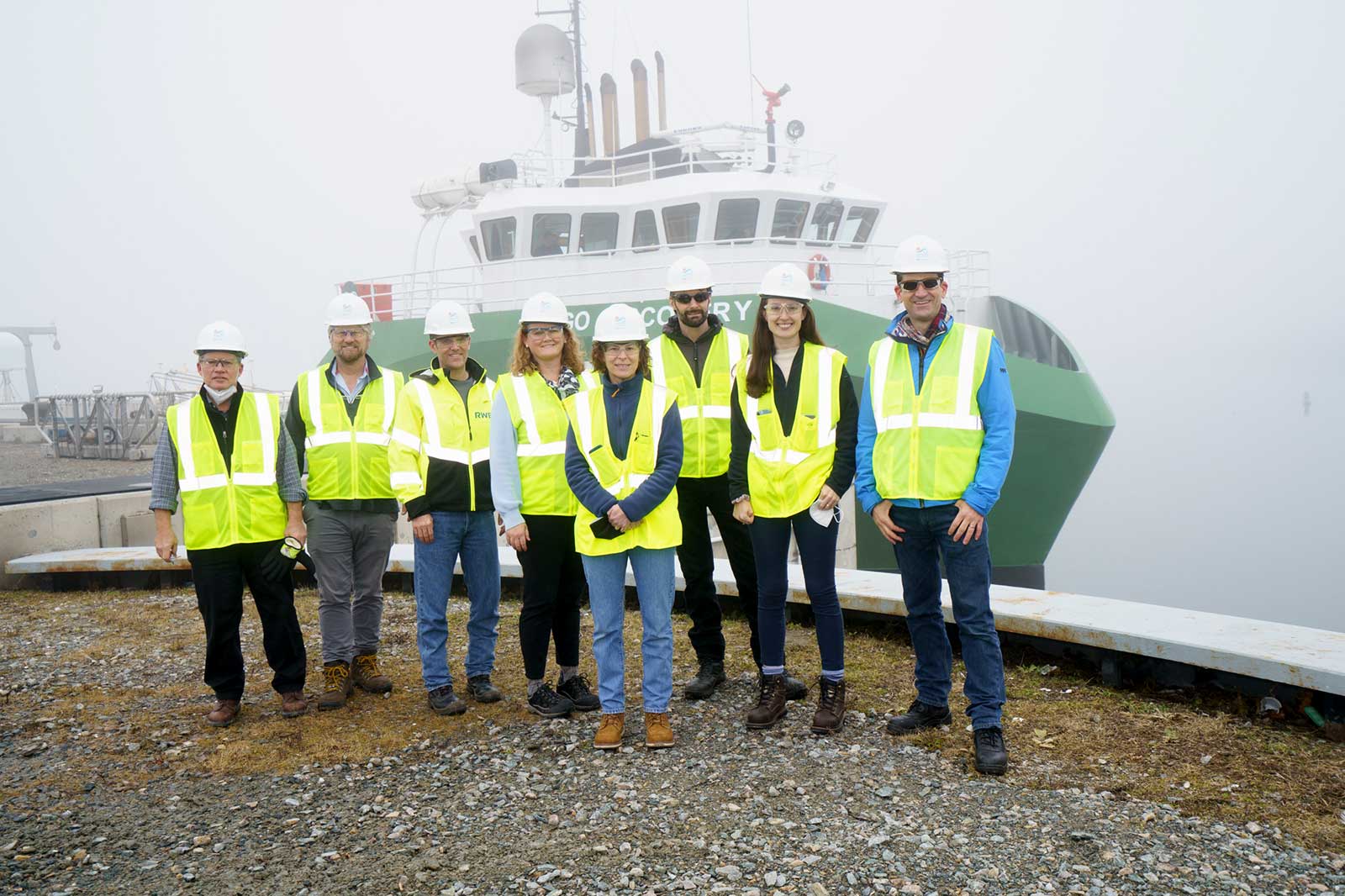 A group in high-visibility jackets and helmets stands on a rocky shore by a ship in fog.
