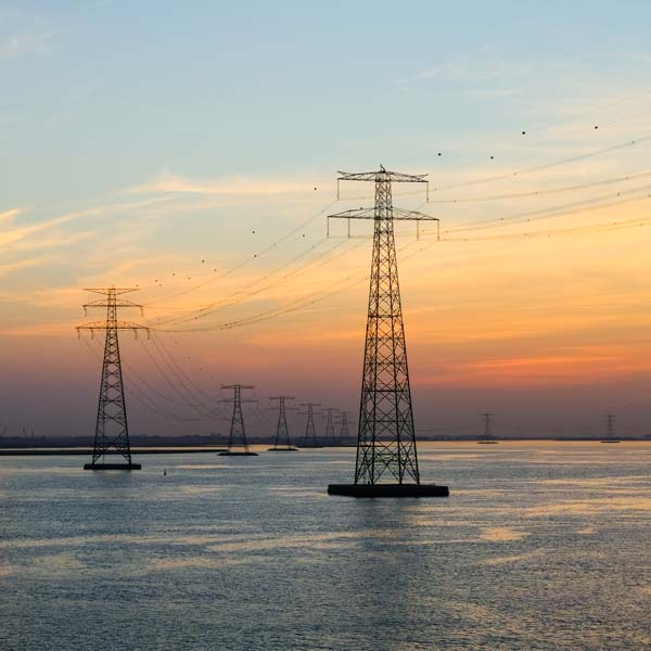 Electricity pylons stand tall above a calm water surface during sunset, with a colourful sky in the background.