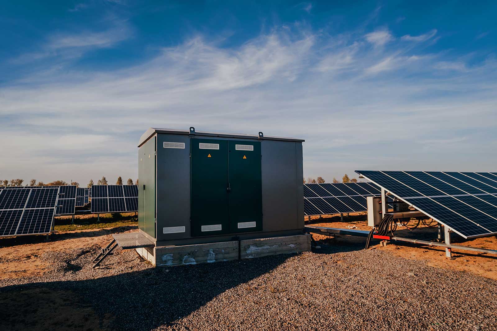 A green electrical cabinet beside solar panels under a clear blue sky, surrounded by gravel and grass.