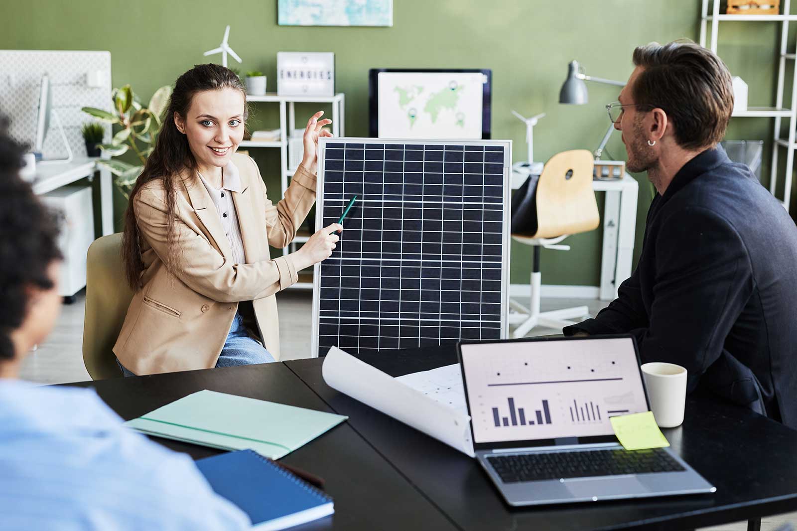 A woman presents a solar panel in a modern office, with colleagues engaged and a laptop showing graphs on the table.