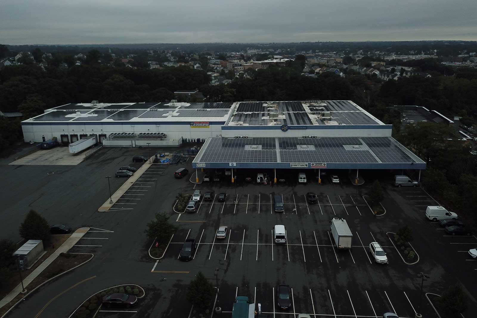 An aerial view of a large commercial building with solar panels, parking areas, and a cloudy sky in the background.