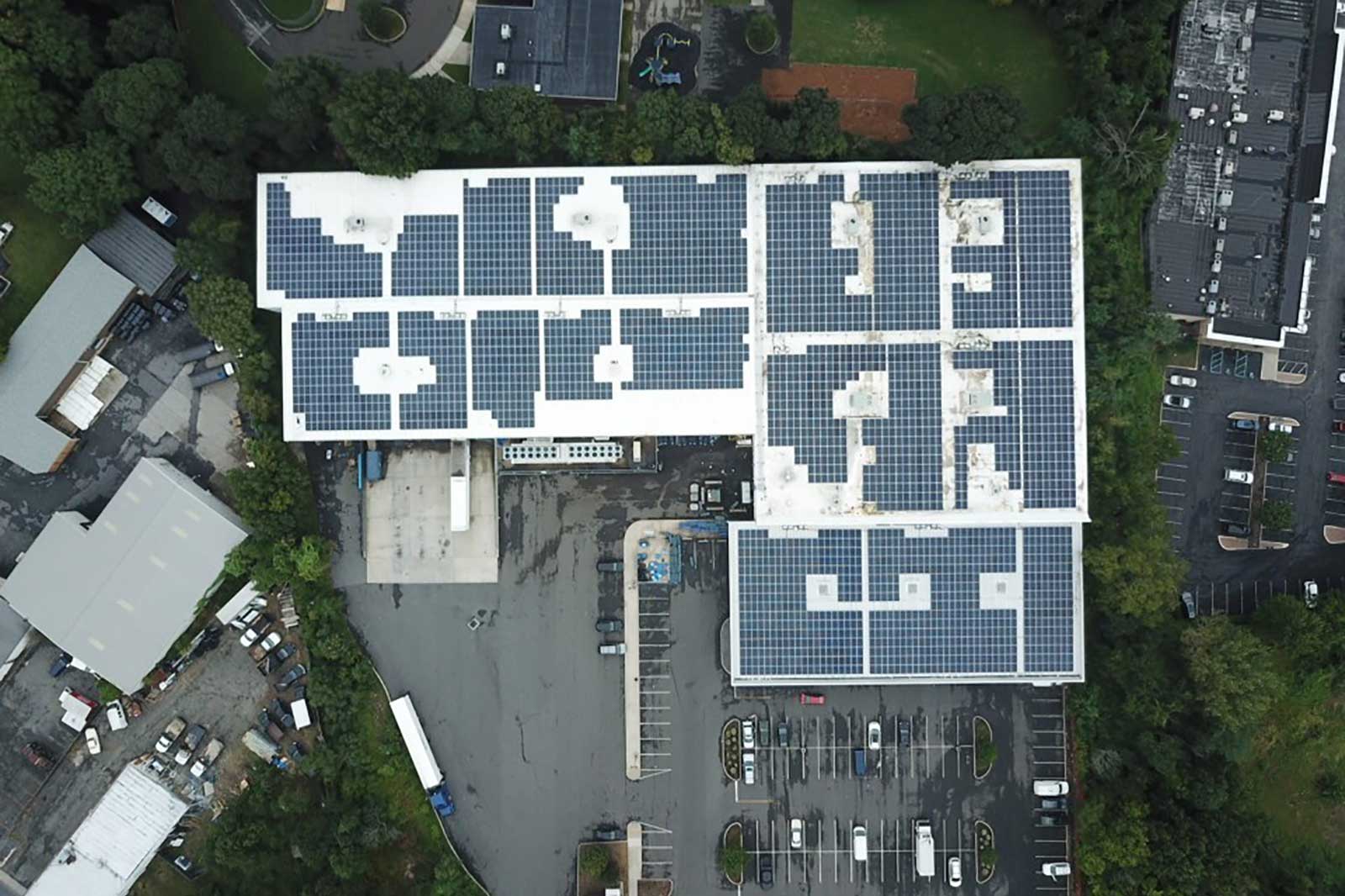 Aerial view of a large building with solar panels on the roof and parking spaces below, surrounded by greenery and other structures.