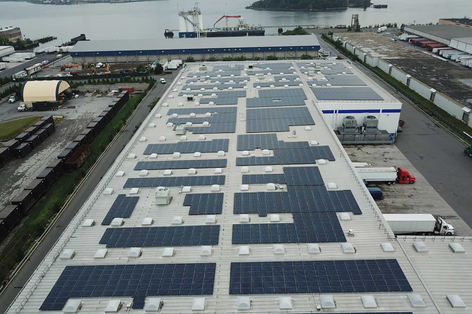 An aerial view of a warehouse roof covered in solar panels, with vehicles and a river in the background.