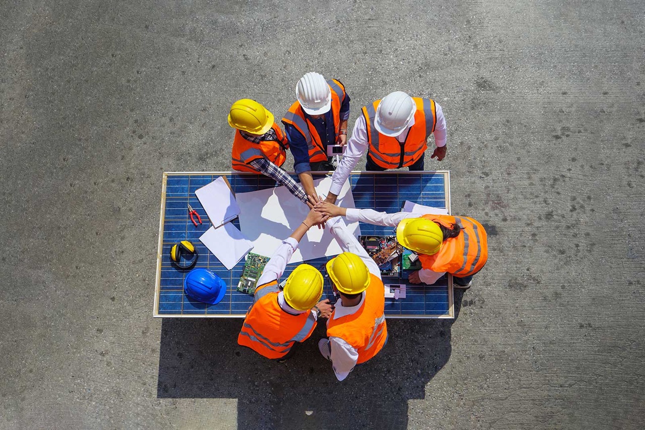 A group of six construction workers in hard hats and high-visibility vests collaborate around a table covered with plans and tools.