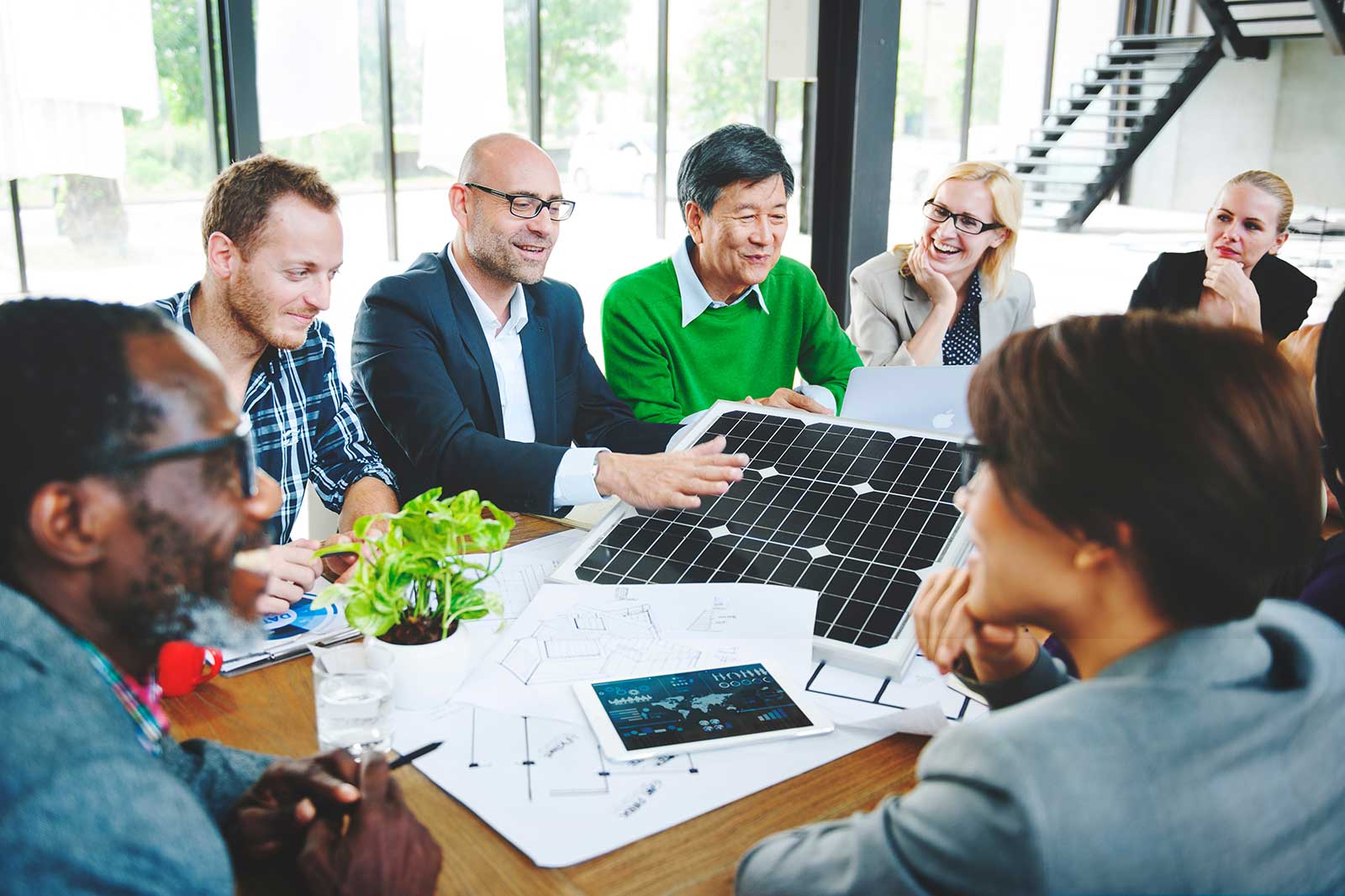 A group discusses a solar panel design in a modern office, with charts and a tablet on the table.