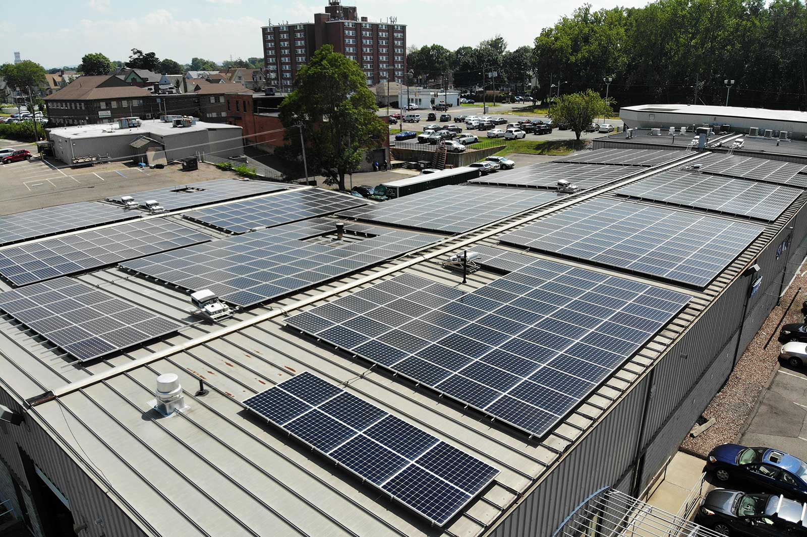 Aerial view of a building's roof covered in solar panels. Nearby, there are cars parked in a lot and a cityscape in the background.