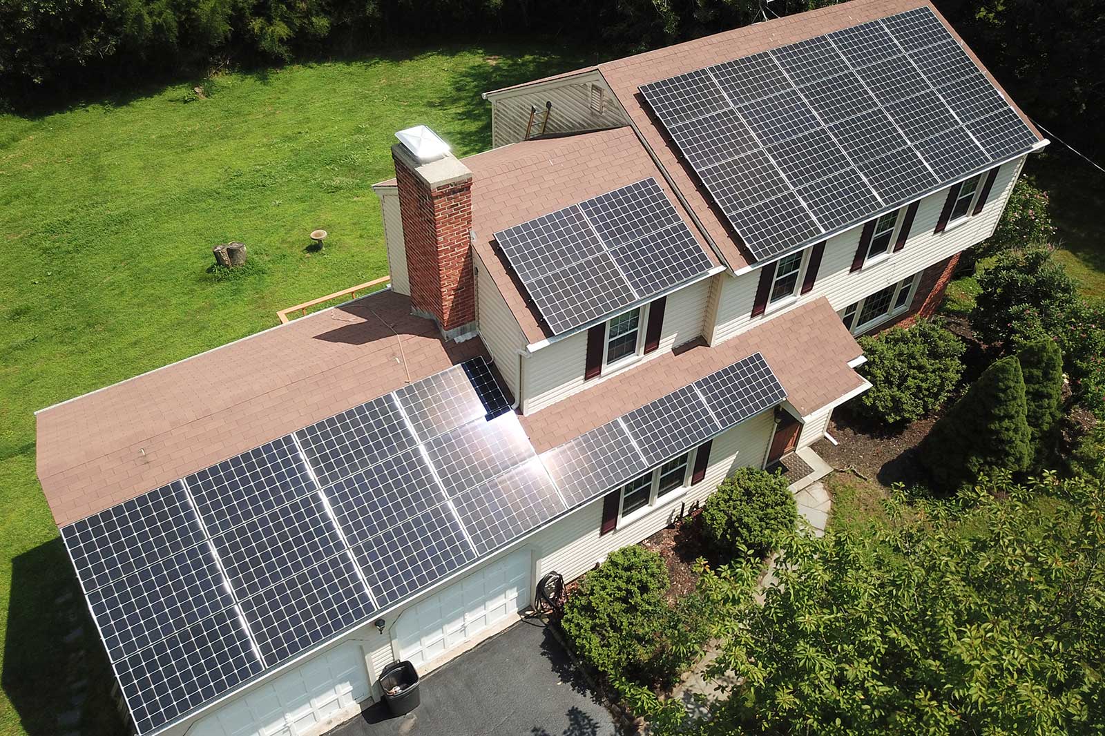 Aerial view of a house with multiple solar panels installed on the roof. Surrounding greenery is visible.