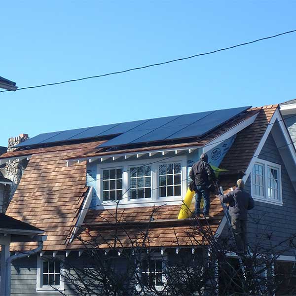Two workers installing solar panels on a sloped roof with wooden shingles under a clear blue sky.