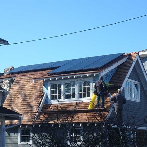 Two workers installing solar panels on a sloped roof with wooden shingles under a clear blue sky.