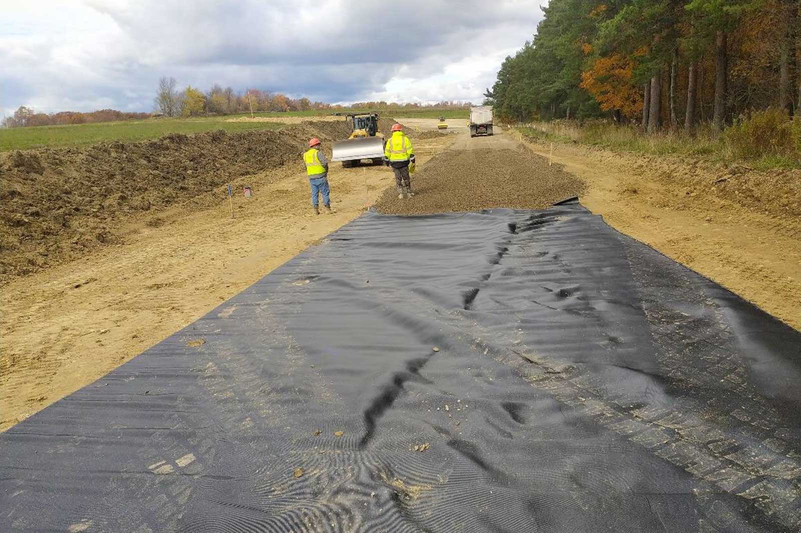 Construction workers lay black geotextile fabric on a dirt road, with machinery and trees in the background.