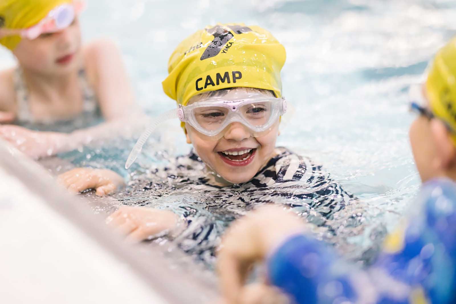 Children swimming in a pool, wearing yellow swim caps. Focus on one child, with hands resting on the pool's edge.