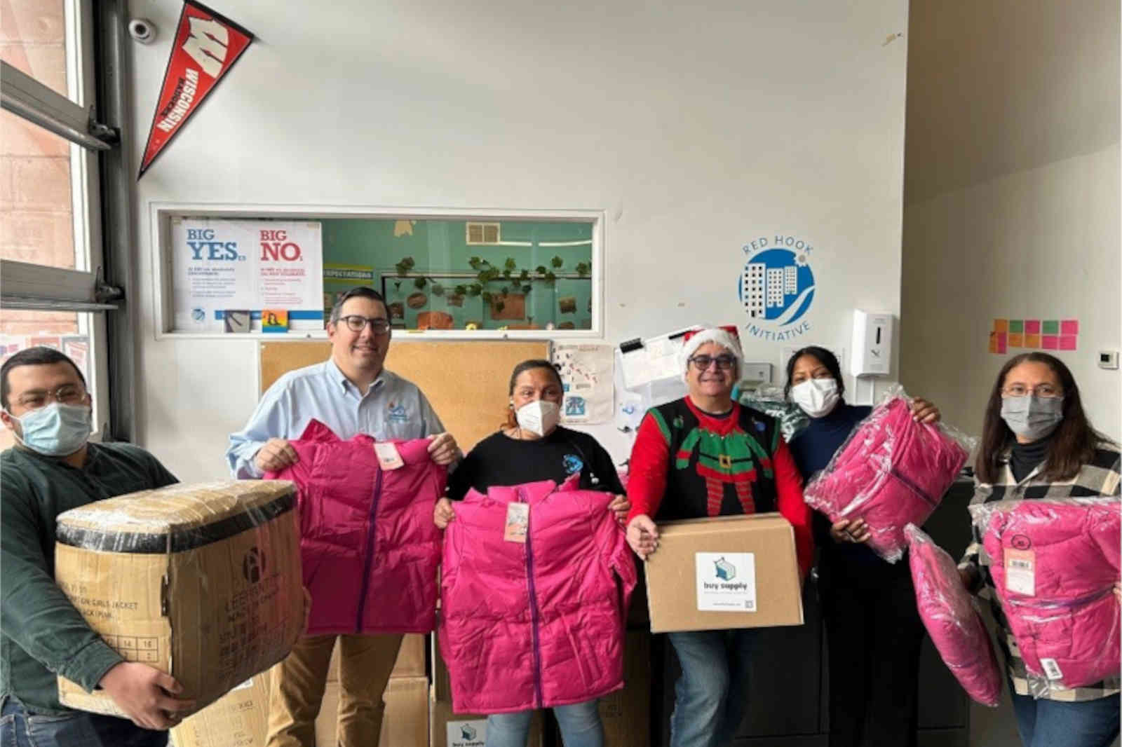 A group of individuals holding pink jackets and a box in a community space with banners and a logo.