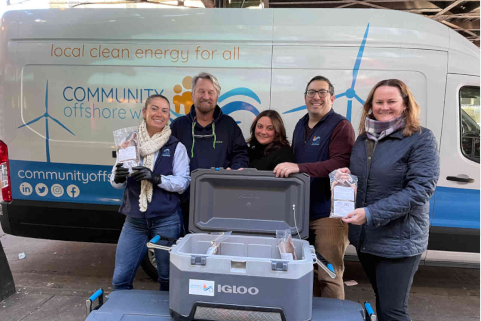 Individuals standing in front of a van promoting community offshore wind energy, holding packages in front of an Igloo cooler.