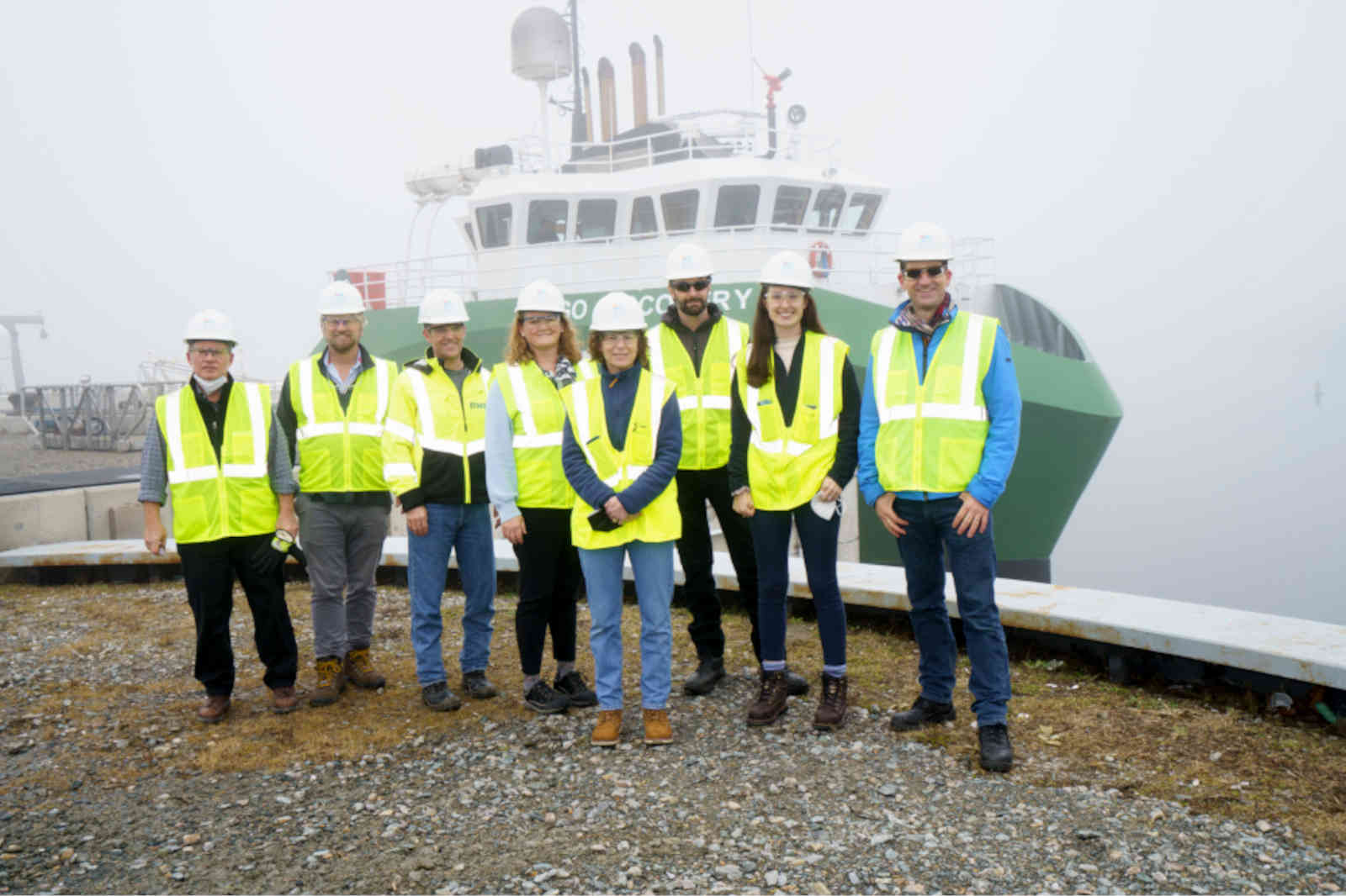 A group of individuals in safety vests and hard hats pose on a foggy dock with a green ship in the background.