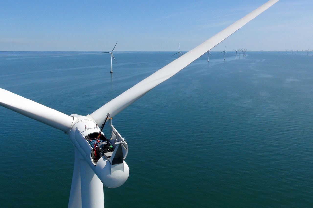A close-up view of a wind turbine on the water, with multiple turbines visible in the background under a clear blue sky.