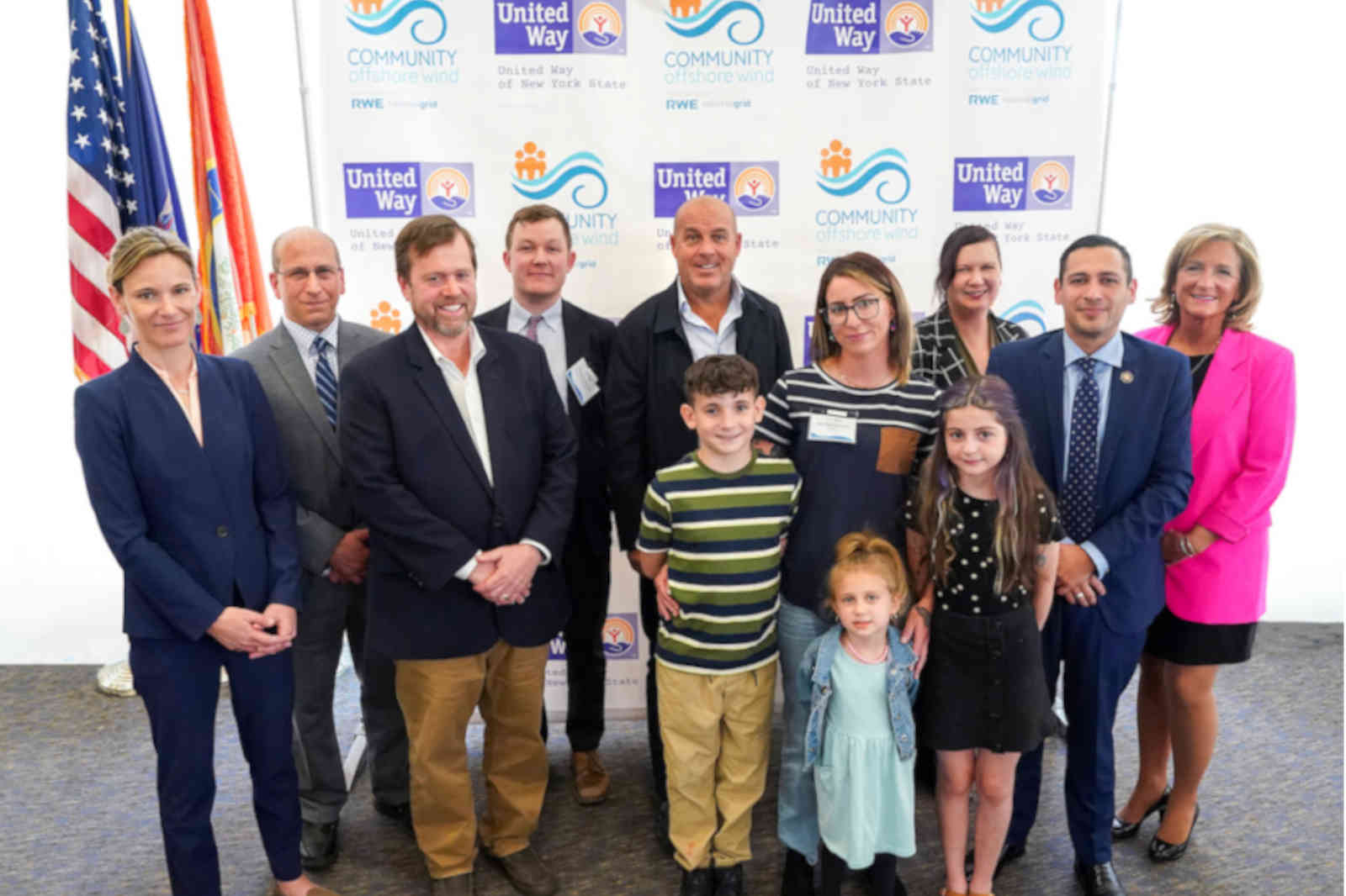 A group photo taken at an event with flags in the background, featuring adults and children posing together.