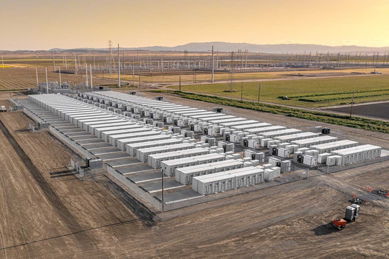 Aerial view of a large energy storage facility with rows of battery containers, surrounded by a vast landscape and power lines.