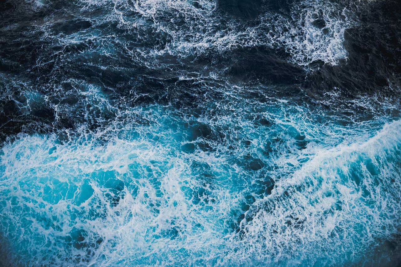 Aerial view of vibrant blue ocean waves crashing against dark waters, creating white foam and intricate patterns.