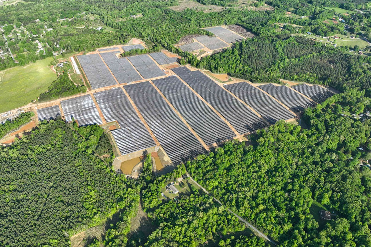 An aerial view of a large solar farm surrounded by green trees and fields, featuring rows of solar panels.