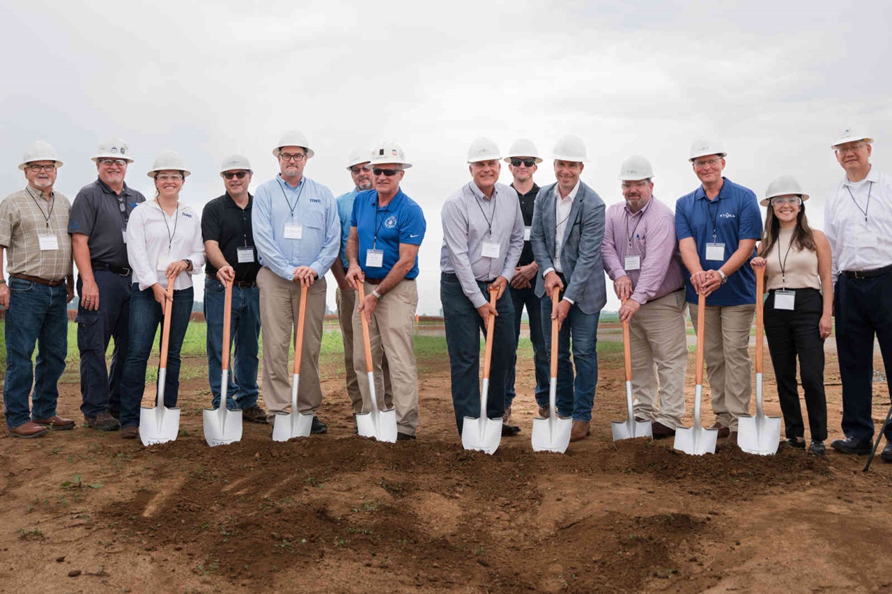 A group of individuals in hard hats holds shovels during a groundbreaking ceremony on a cloudy day. The group stands on soil.