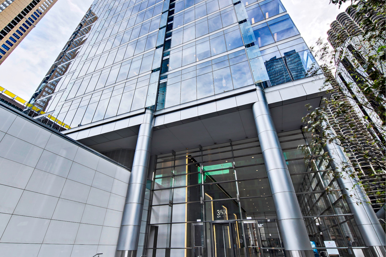 A modern glass-fronted building with a sleek entrance, reflecting nearby structures and a cloudy sky.