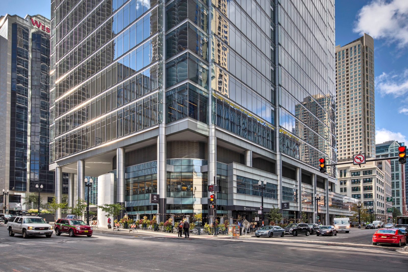 A modern glass skyscraper reflects the city skyline, with busy streets and traffic lights in the foreground.